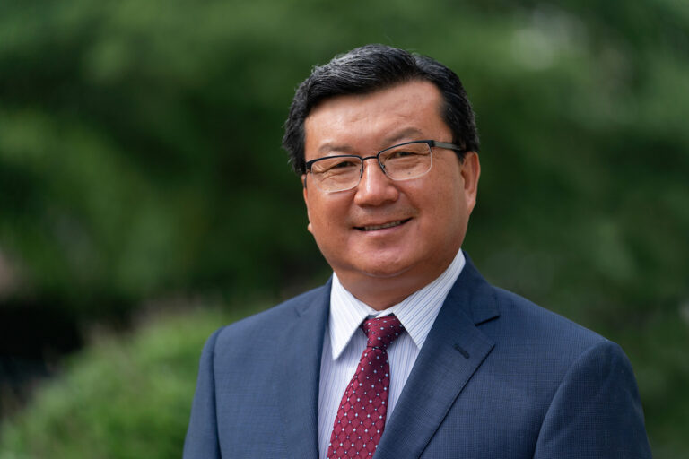 a smiling man in a maroon tie and blue suit in an outdoor setting, Wenwei Xu