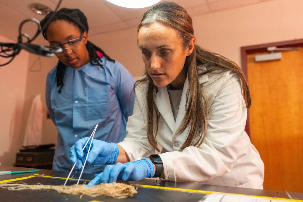 A woman in a white lab coat is holding tongs working with plant matter. A woman in blue scrubs stands beside her watching.