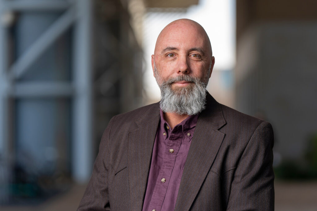 Man with grey beard, slightly smiling in jacket and collared shirt. Blurred architectural background.