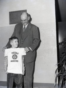 Man in a suit holds up a Texas Aggies shirt up to a kids body
